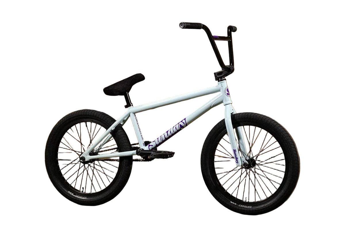 A white BMX bike with black handlebars, seat, and tires, featuring the Sunday brand logo on the frame, set against a brown cardboard backdrop with the Sunday logo printed on the wall.