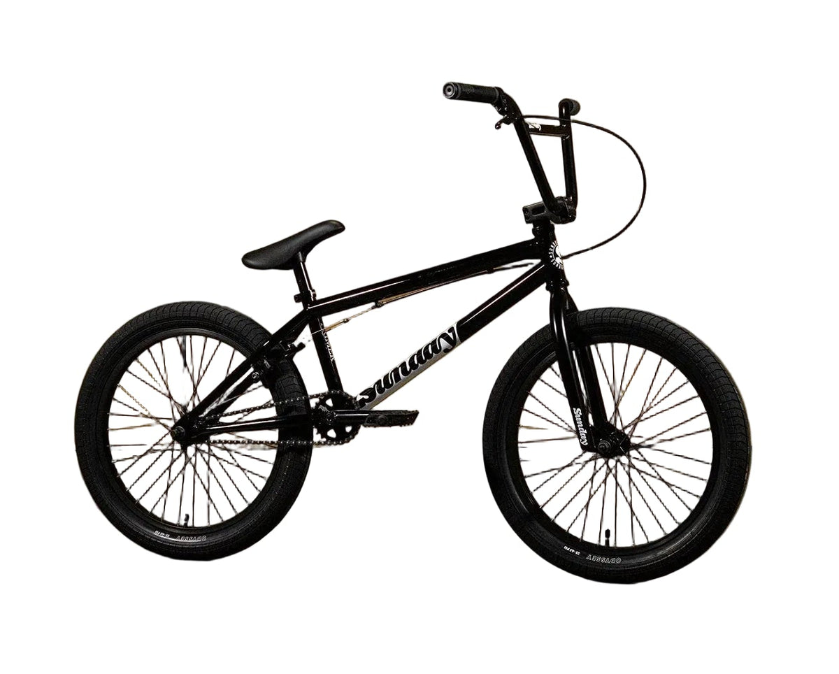 A black Sunday BMX bike with knobby tires and a step-through frame, positioned in front of a yellow-brown paneled wall with the brand name 'Sunday' printed on it.