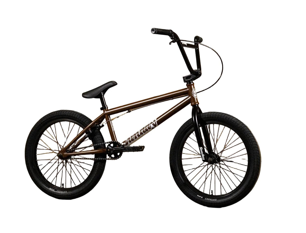 A BMX bicycle with a metallic bronze frame and black components, positioned in profile against a yellow wall with 'Sunday' branding.