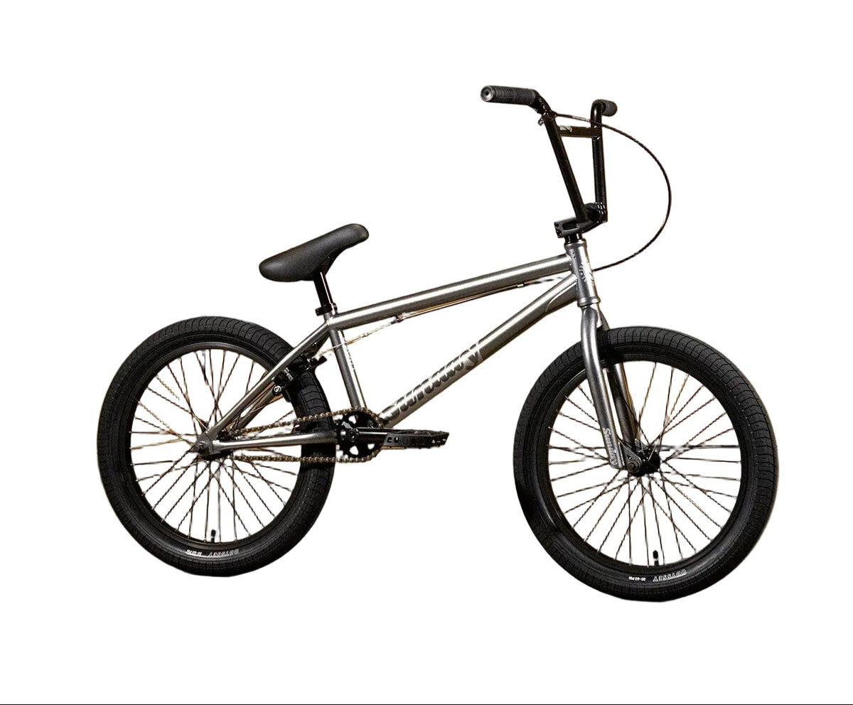 A silver BMX bicycle with black tires, handlebars, and saddle, displayed against a yellow wall with the brand name 'Sunday' printed on it.