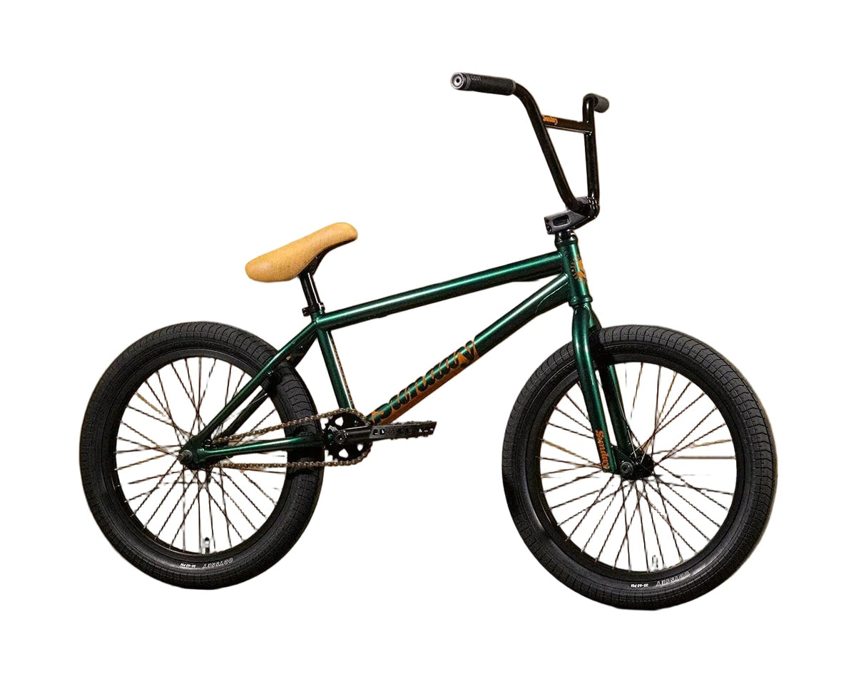 A dark green BMX bike with a tan seat and black handlebars, positioned in profile against a yellow cardboard wall with the brand name 'Sunday' printed on it.