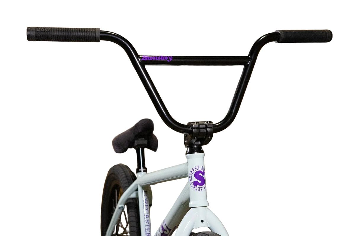A BMX bicycle with a white frame and black handlebars, featuring purple 'Sunday' branding on the frame and top tube, set against a background of stacked cardboard boxes with the word 'Sunday' printed in black.