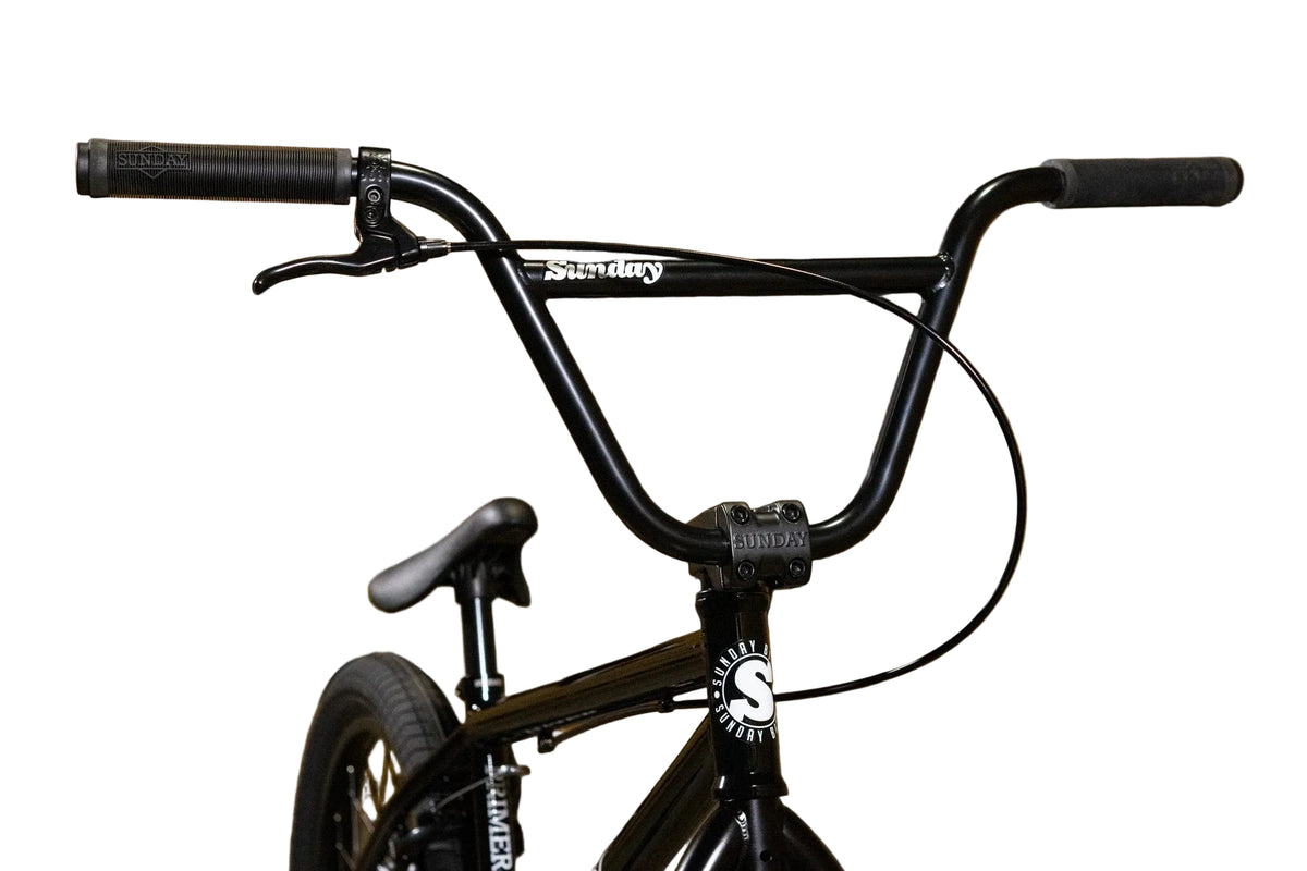 Close-up of a black BMX bike handlebars and frame with a white Sunday brand logo, set against a yellow background with large Sunday text.